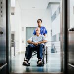 Nurse and Senior Patient in Wheelchair entering a hospital elevator.
