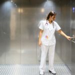 Full body woman in white medical uniform pushing button inside elevator while working in hospital